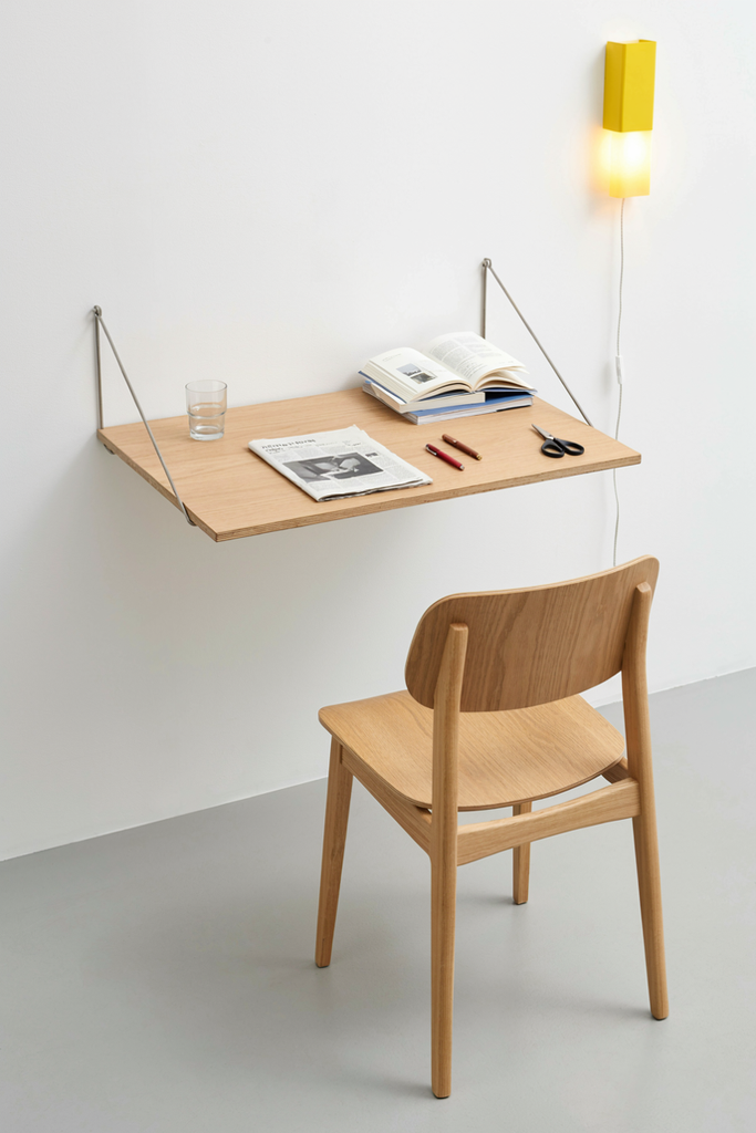 Wall-mounted fold-down wooden desk with a wooden chair and a yellow wall lamp in a small studio apartment